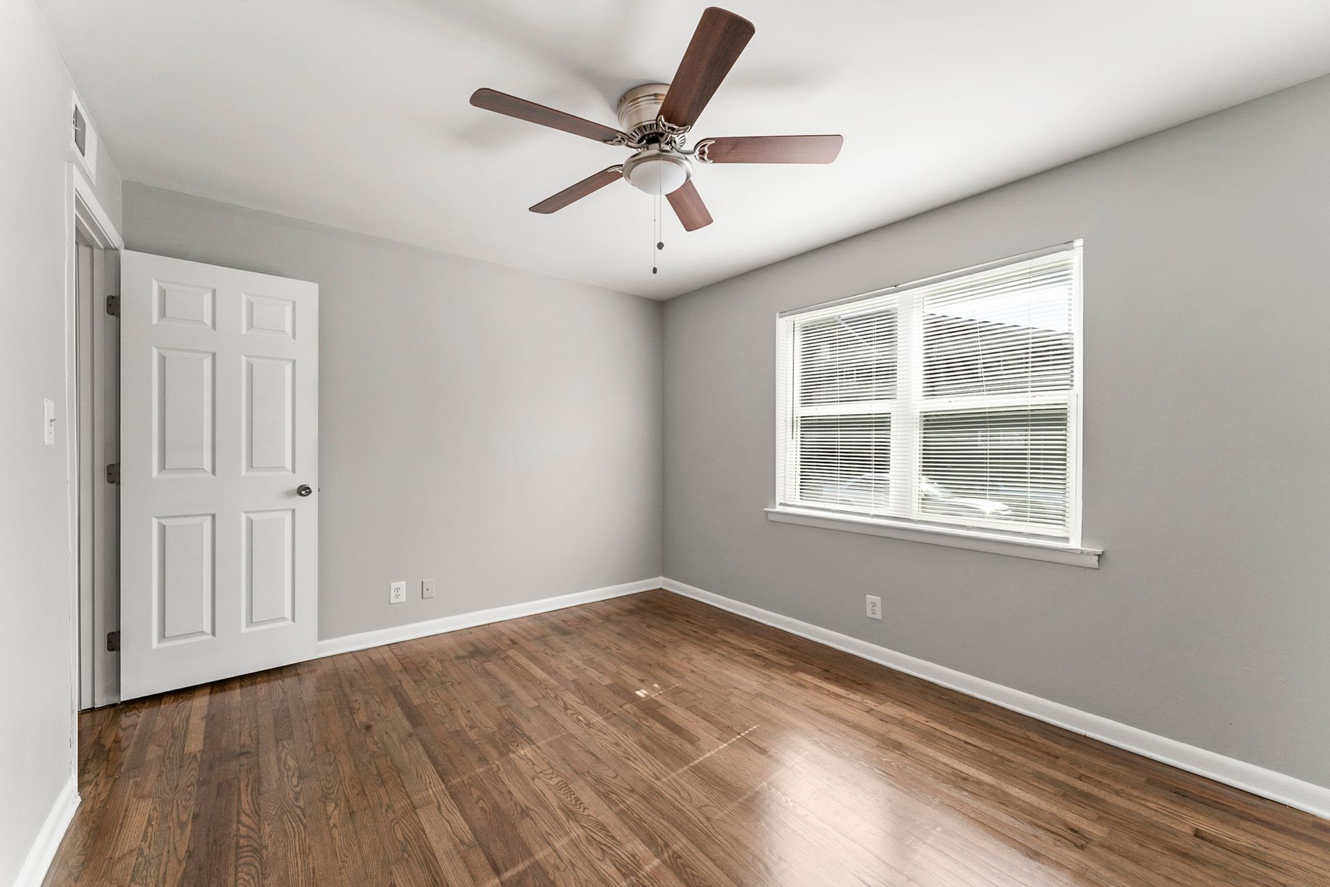 Empty gray bedroom with wood floor, white closet doors, ceiling fan, and window blinds.