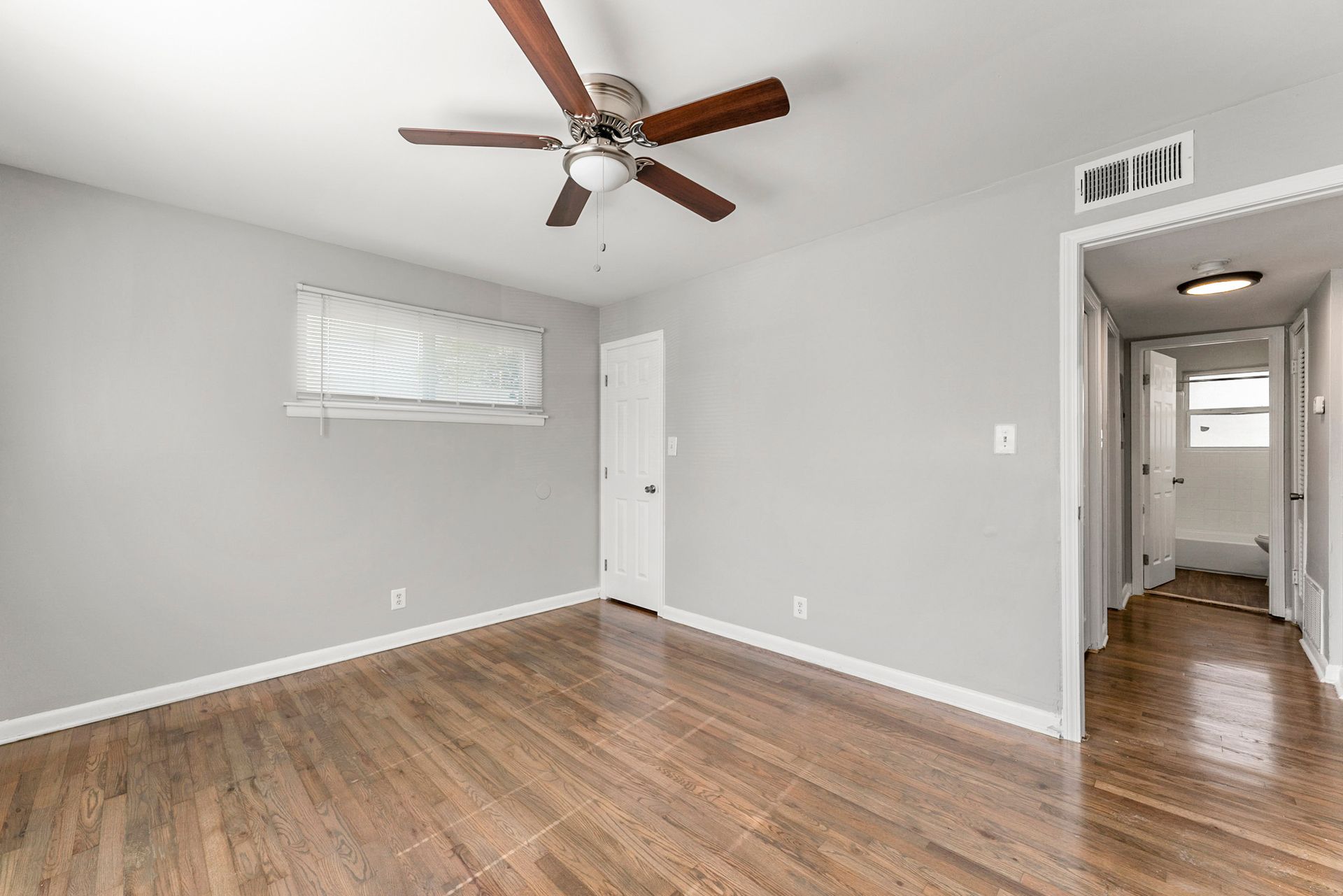 Empty bedroom with gray walls, wood floor, ceiling fan, small window, and open doorway to a hallway
