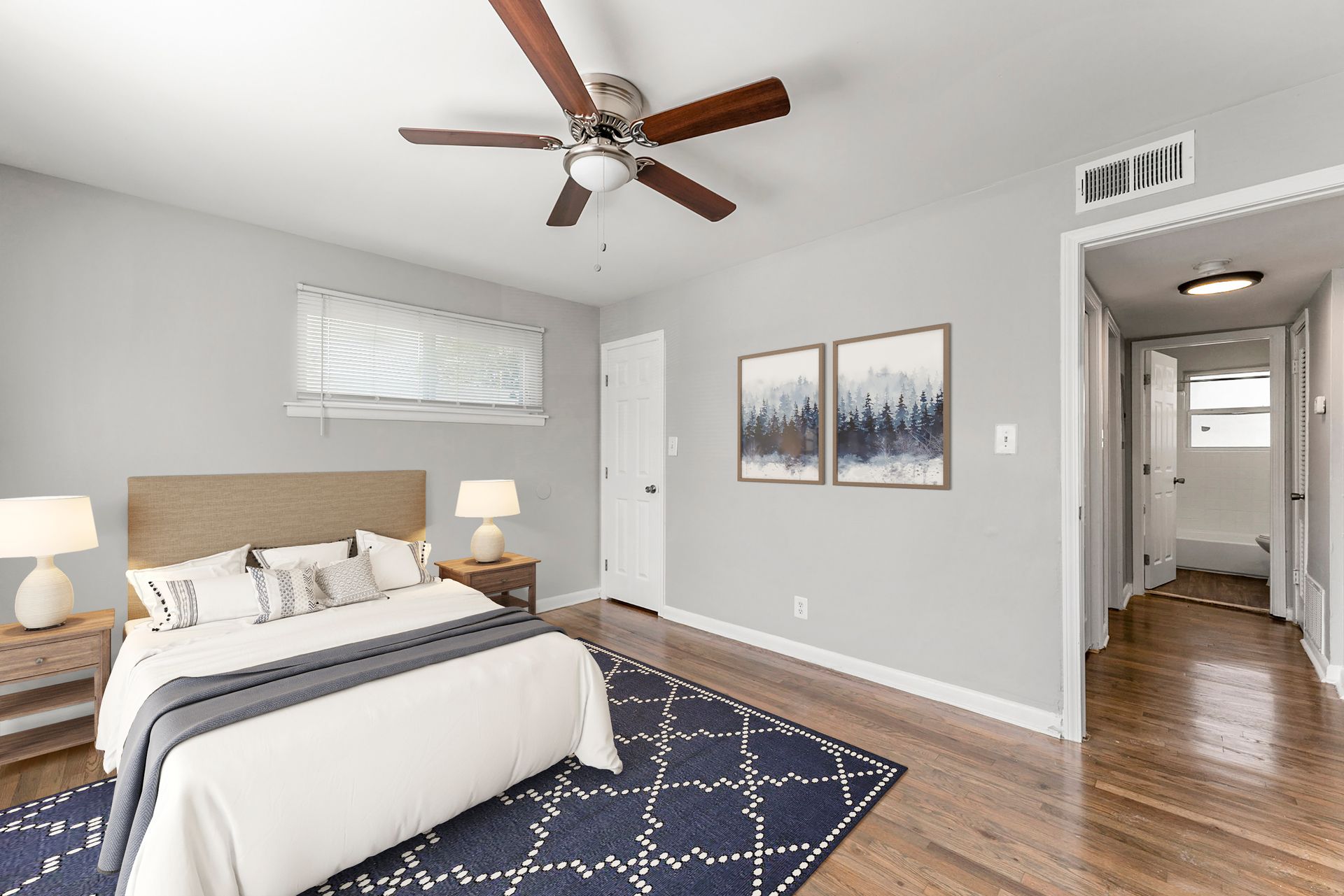 Bright bedroom with queen bed, navy rug, ceiling fan, nightstands, and hallway visible through open door.