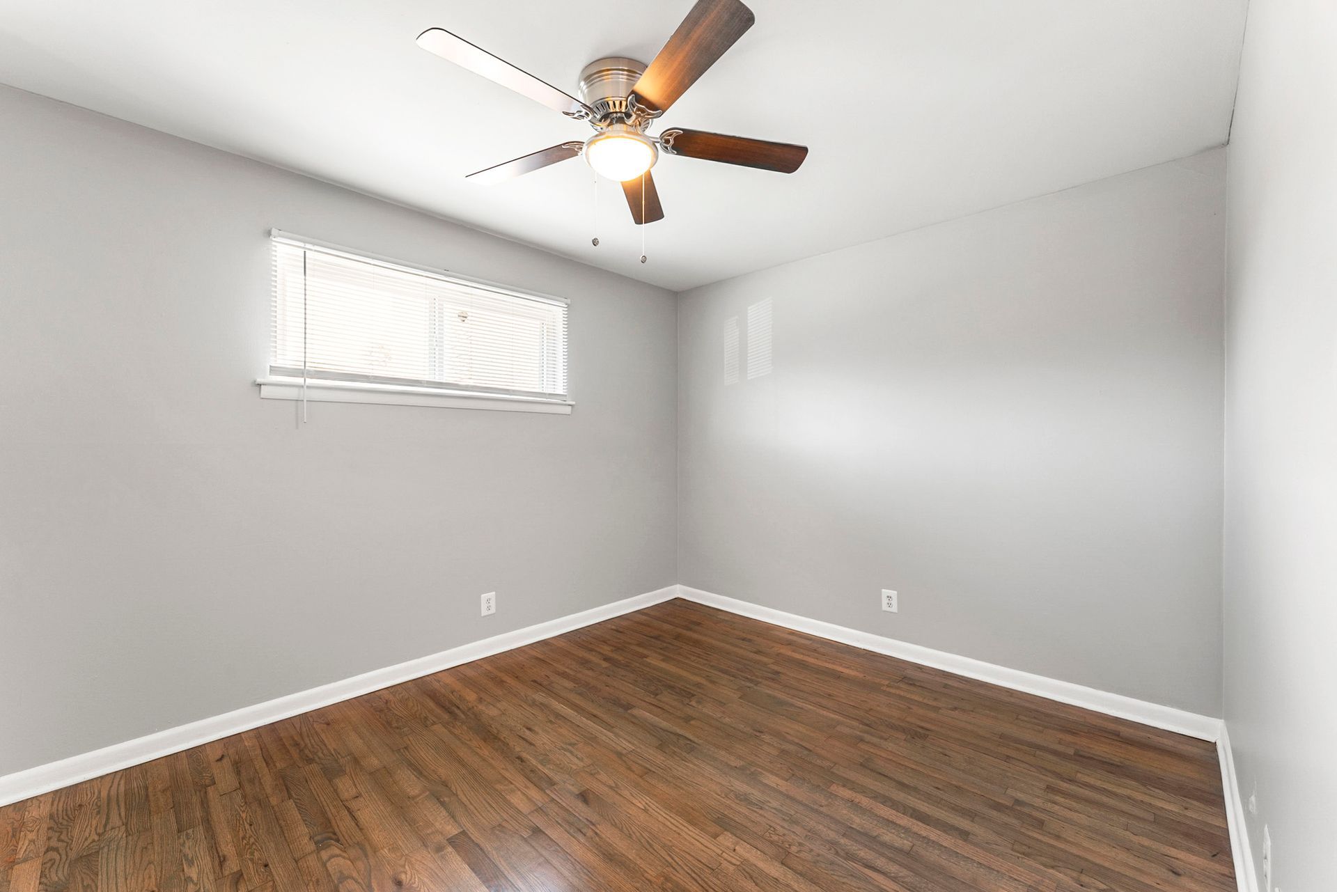 Empty gray bedroom with wood floor, white trim, a window, and a ceiling fan with light.
