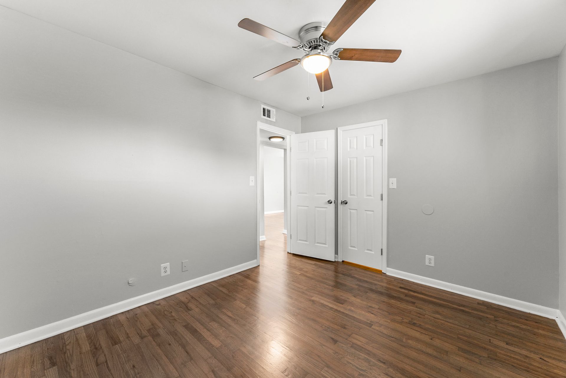 Empty bedroom with gray walls, hardwood floor, white closet doors, and a ceiling fan on