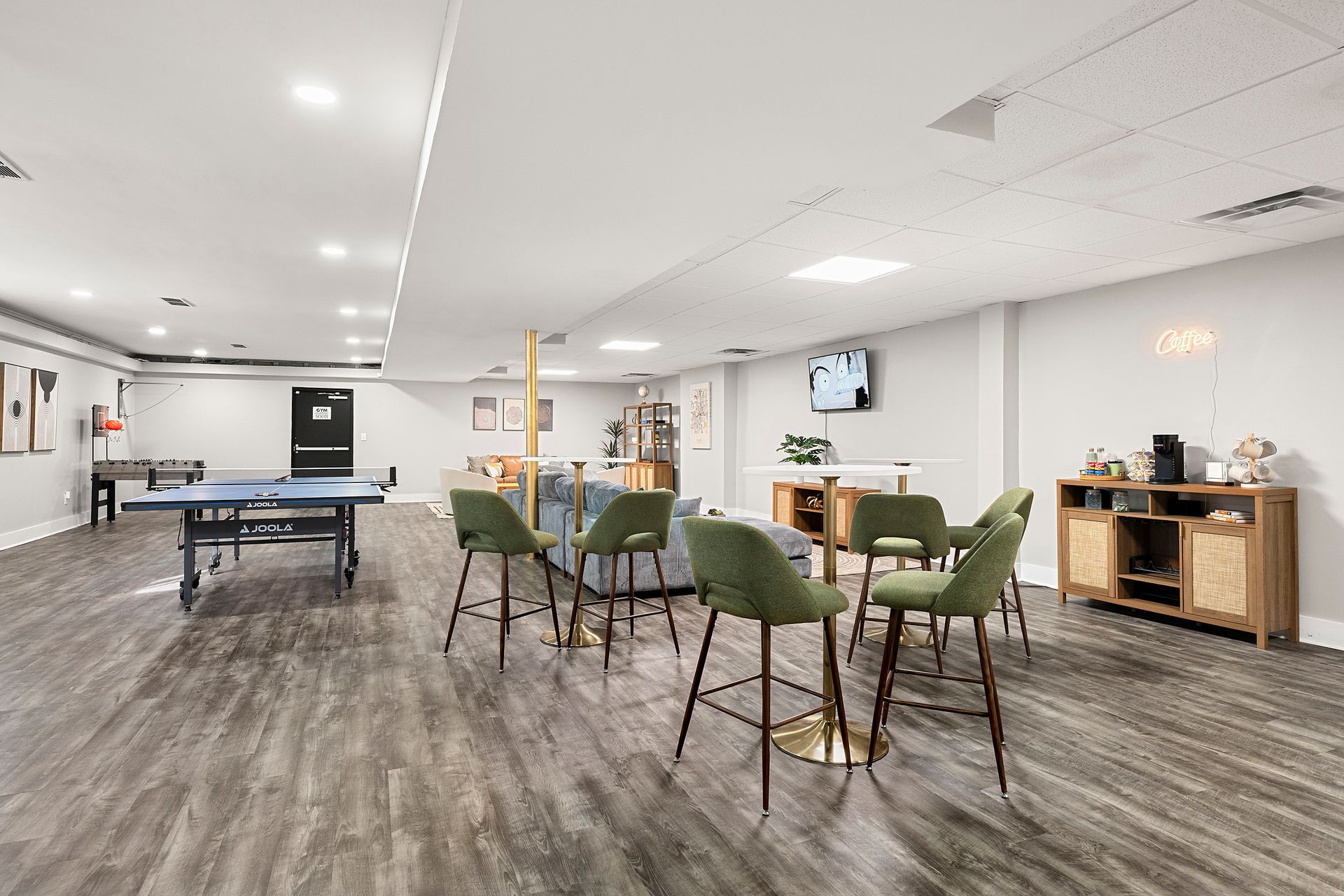 Spacious rec room with tables, green chairs, TV, and wood-look flooring in a bright basement setting
