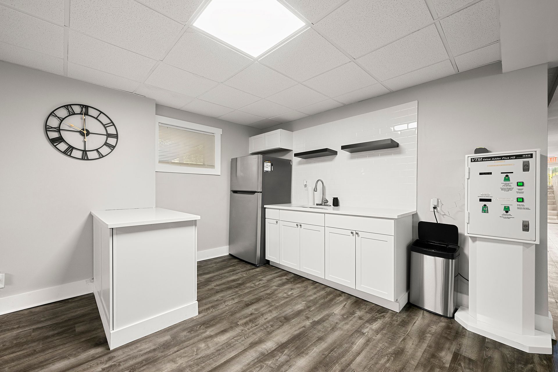 Small break room with white cabinets, sink, fridge, trash can, wall clock, and wood-pattern flooring.