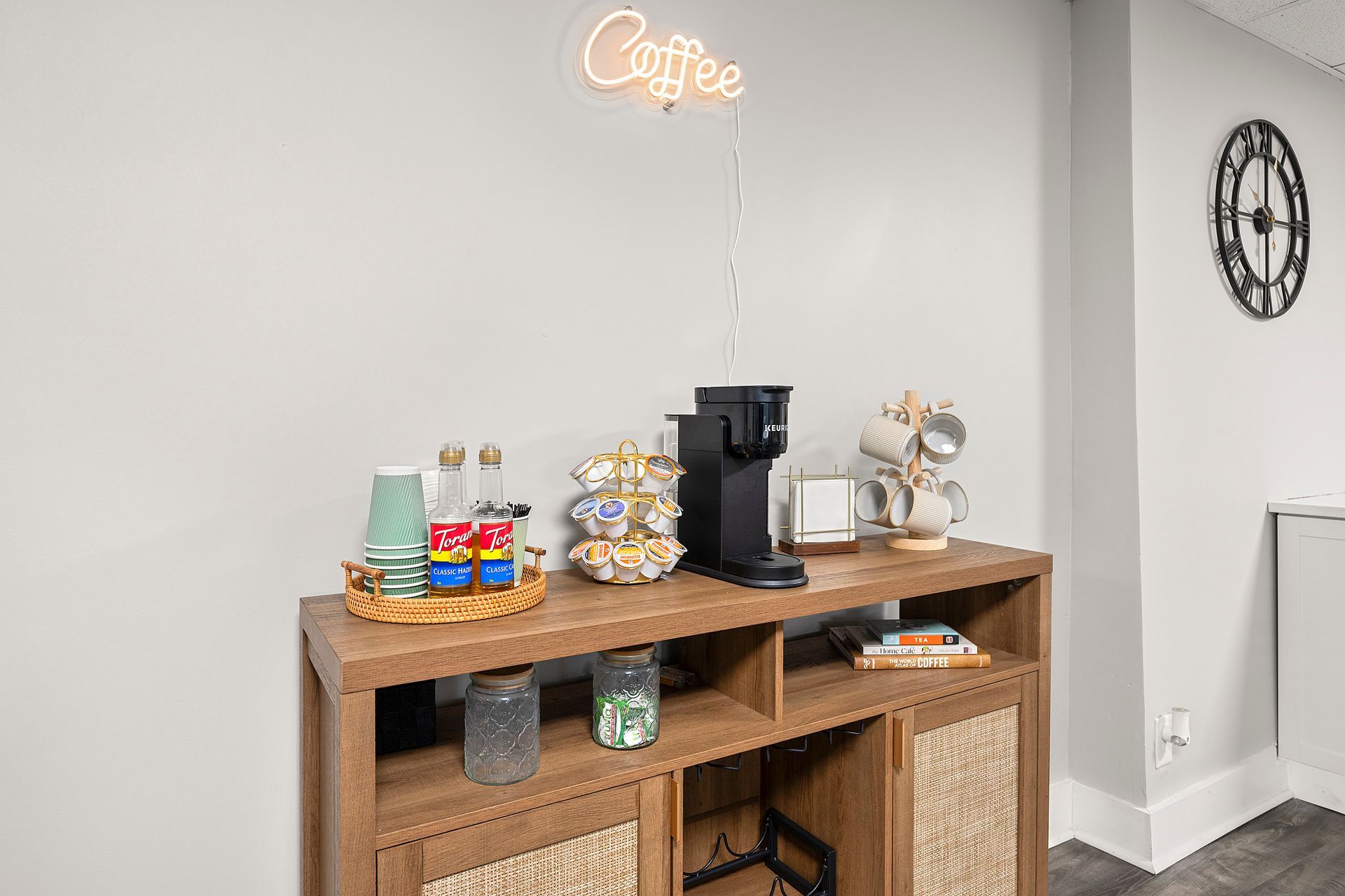 Coffee station with wooden cabinet, mugs, snacks, and espresso machine against a light gray wall.