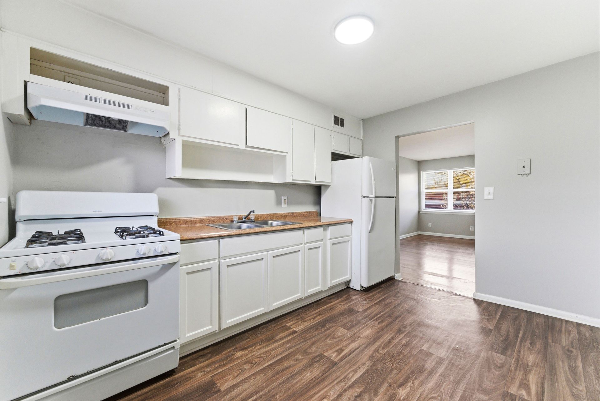 Bright white kitchen with stove, cabinets, refrigerator, and wood-look flooring leading to a doorway