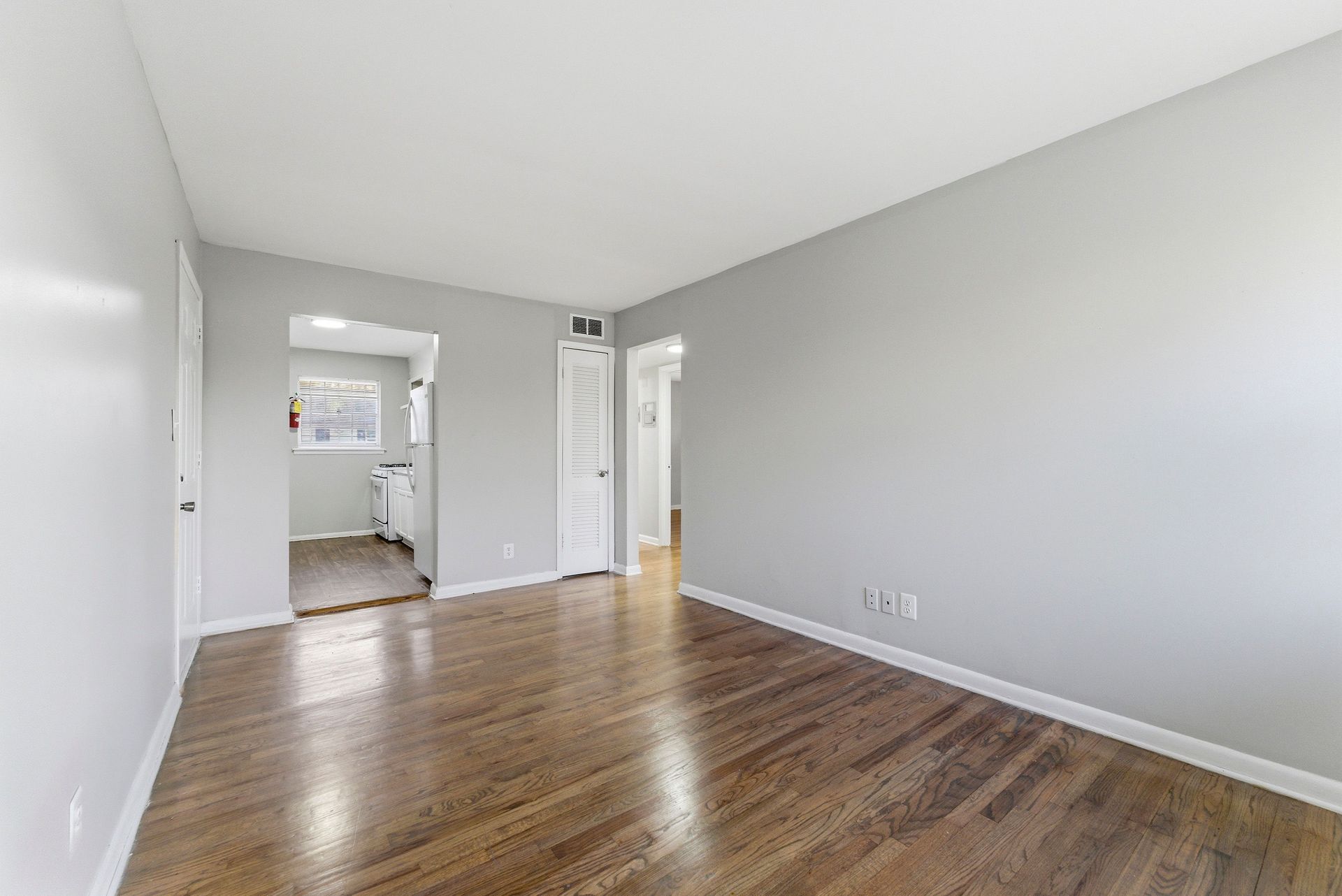 Empty living room with gray walls, hardwood floors, and a view into a kitchen area.