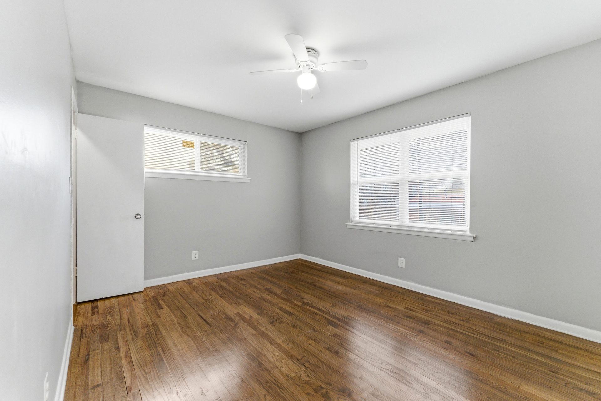 Empty gray bedroom with hardwood floors, two windows, and a ceiling fan light.