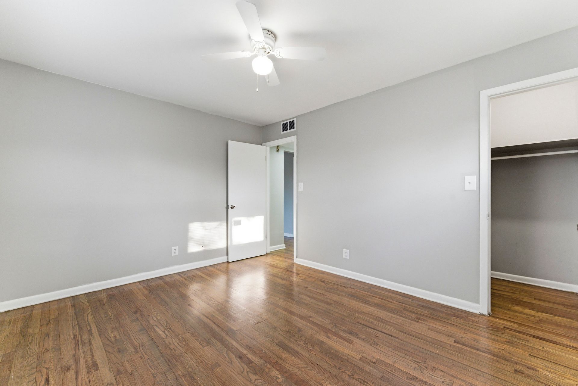 Empty gray bedroom with hardwood floor, ceiling fan, open door, and closet.