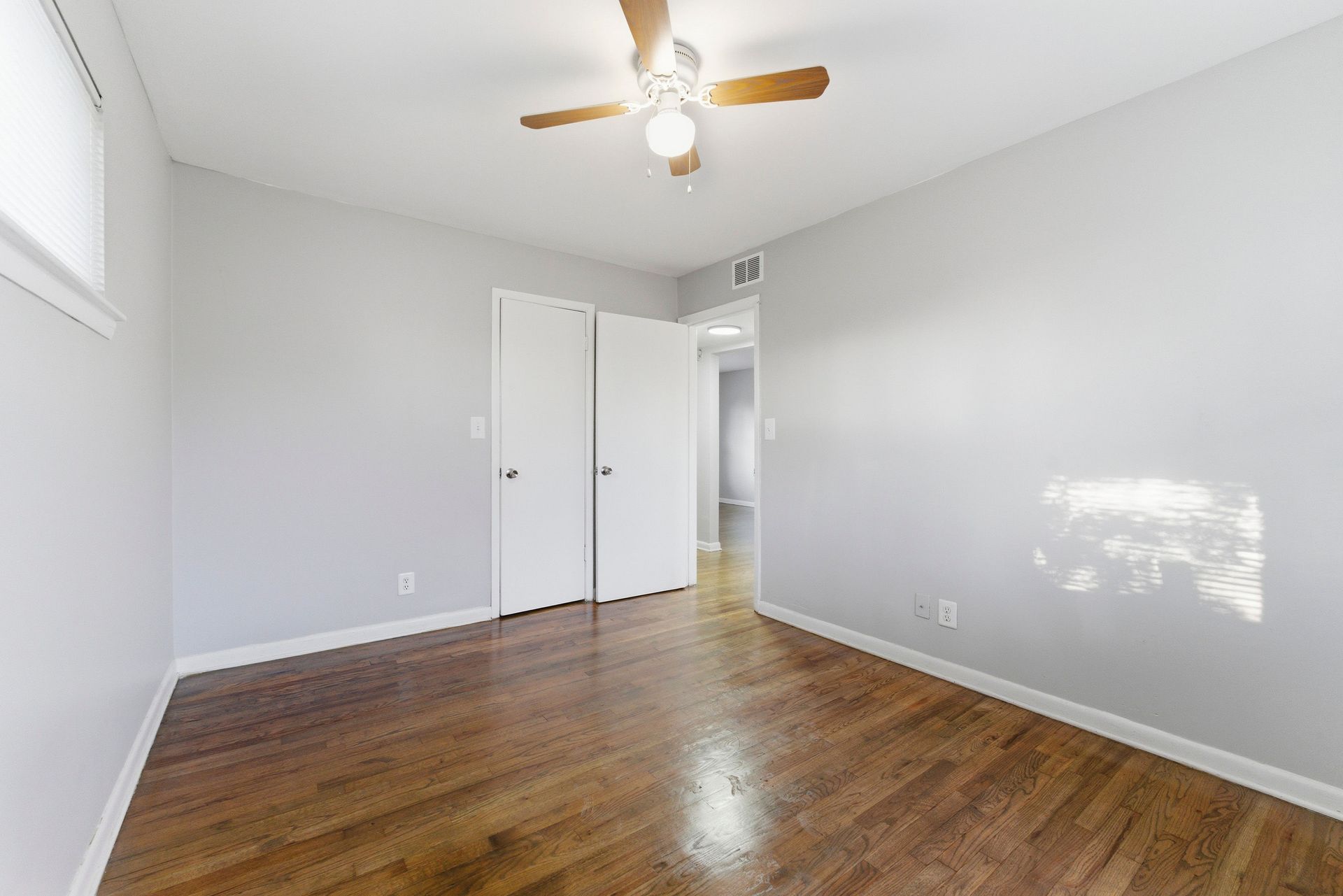 Empty light-gray bedroom with wood floor, white closet doors, ceiling fan, and sunlight on the wall.