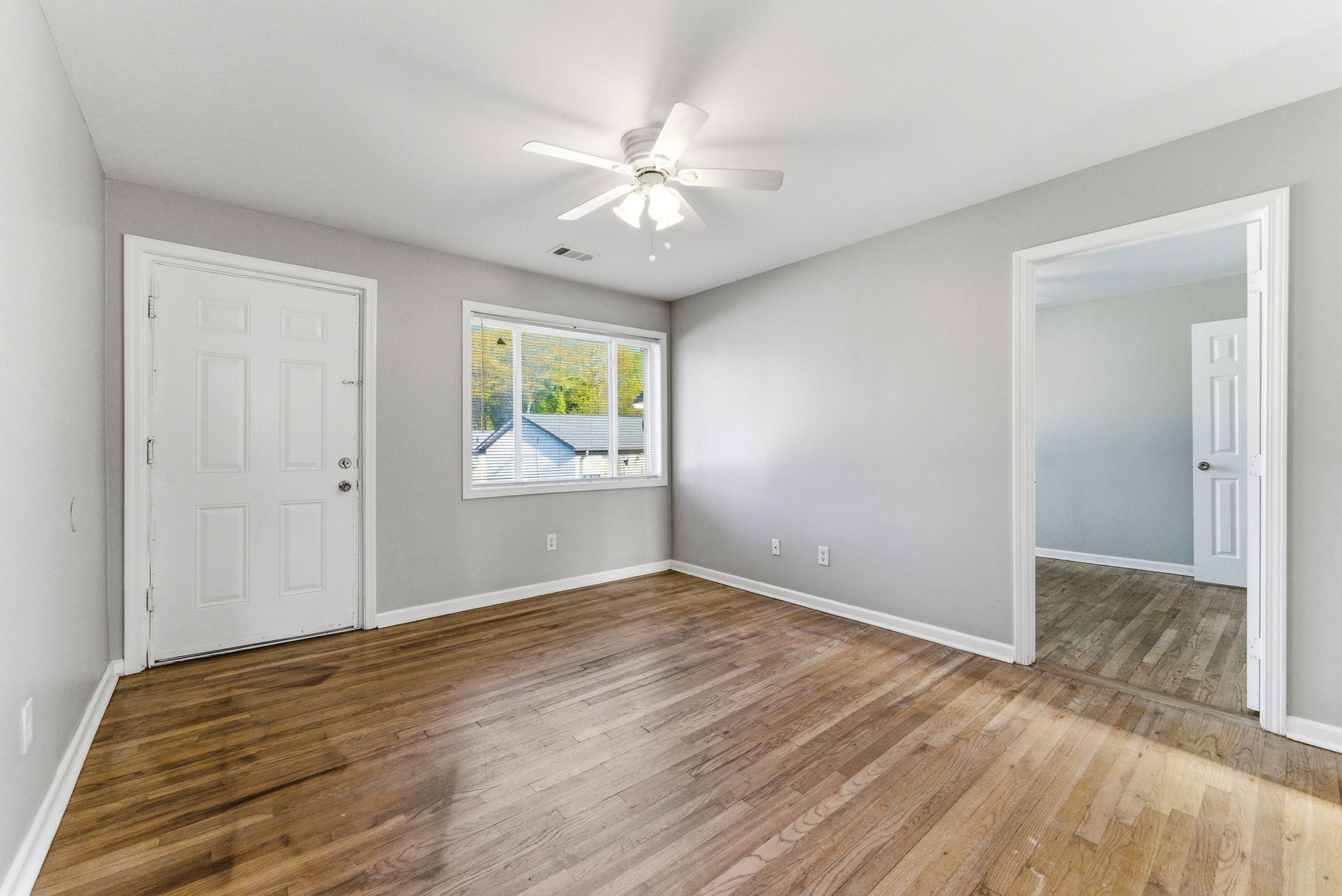 Empty gray bedroom with wood floors, ceiling fan, window, and open doorway to adjoining room