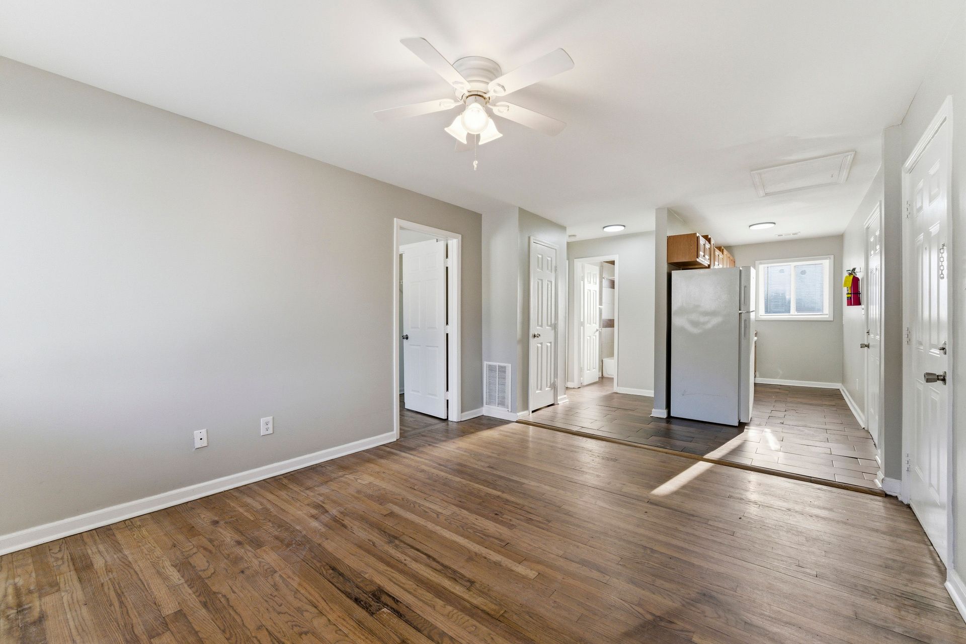Empty living room with wood floors, white walls, ceiling fan, and kitchen area in the back.