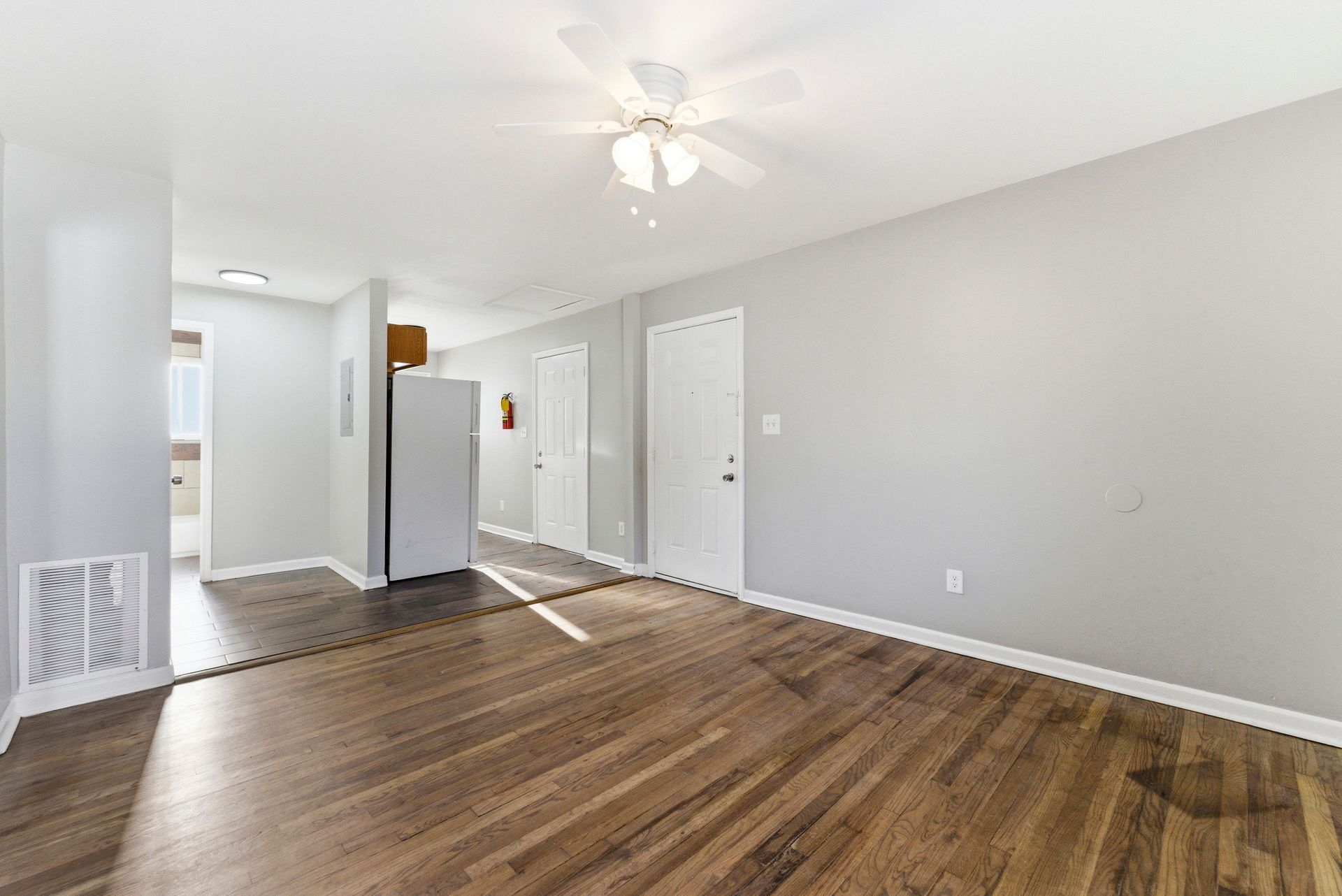 Empty living room with wood floors, white walls, ceiling fan, and kitchen in the background