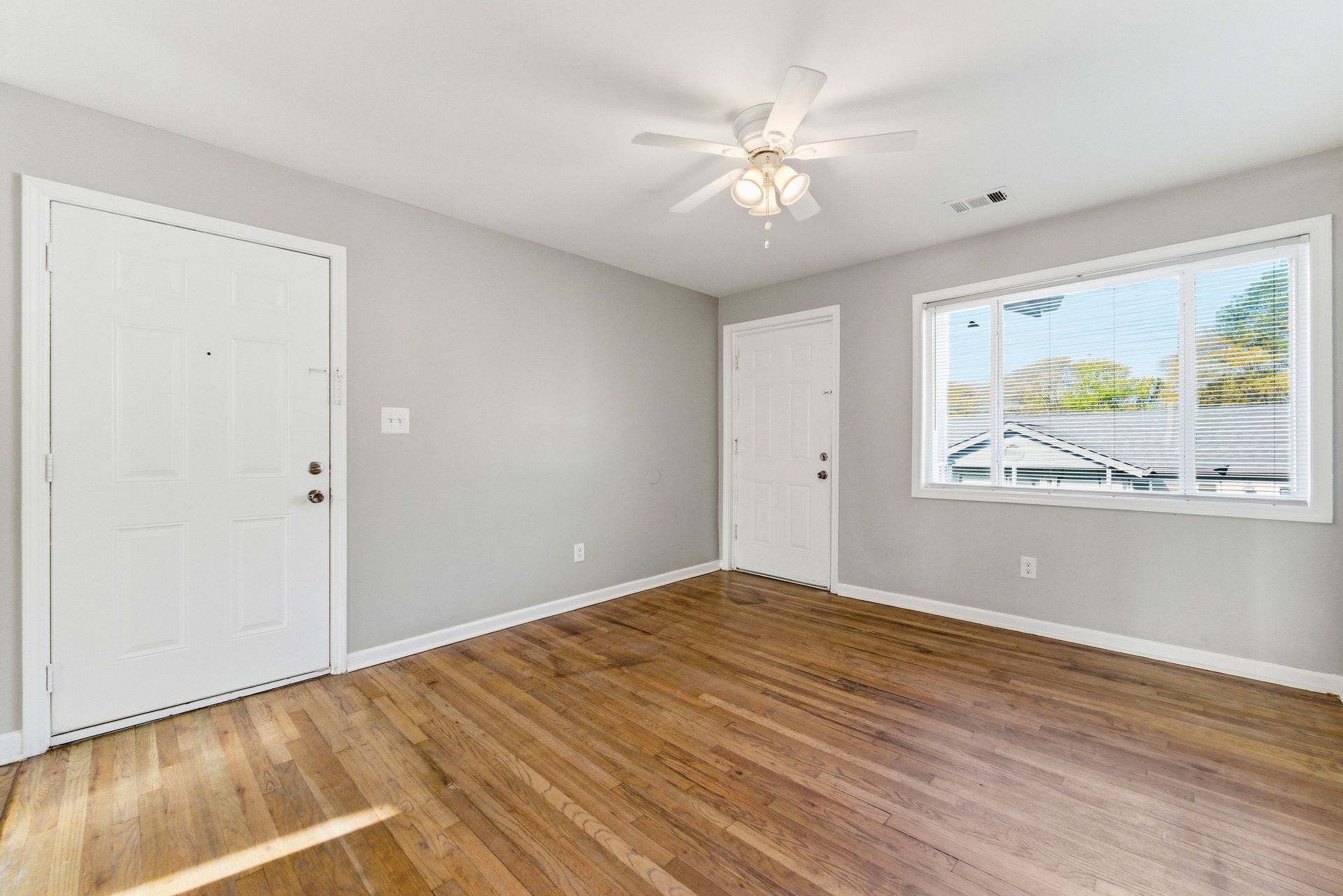 Empty bedroom with light gray walls, hardwood floors, white closet doors, and a window with blinds.