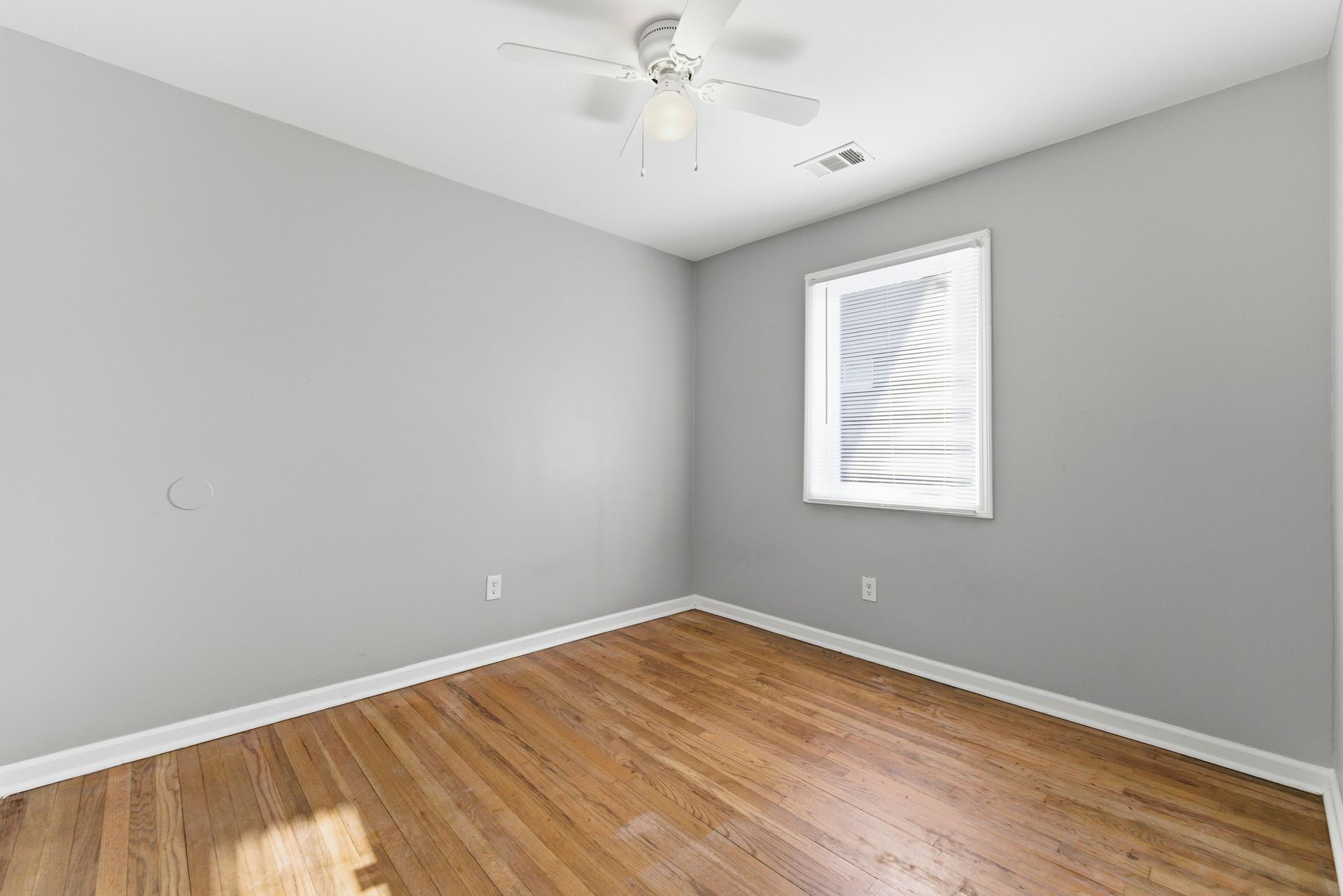 Empty gray room with hardwood floors, a ceiling fan, and a small window with blinds.