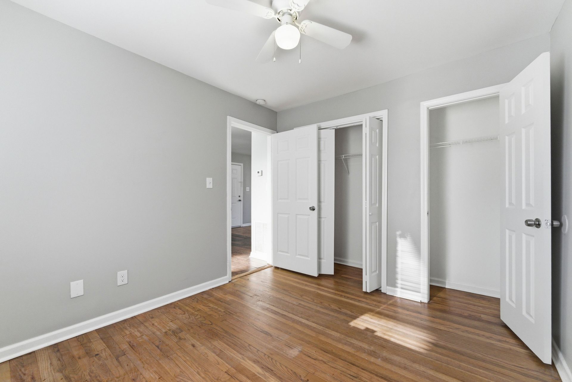 Empty gray bedroom with hardwood floors, ceiling fan, and two open white doors to a hallway and closet.