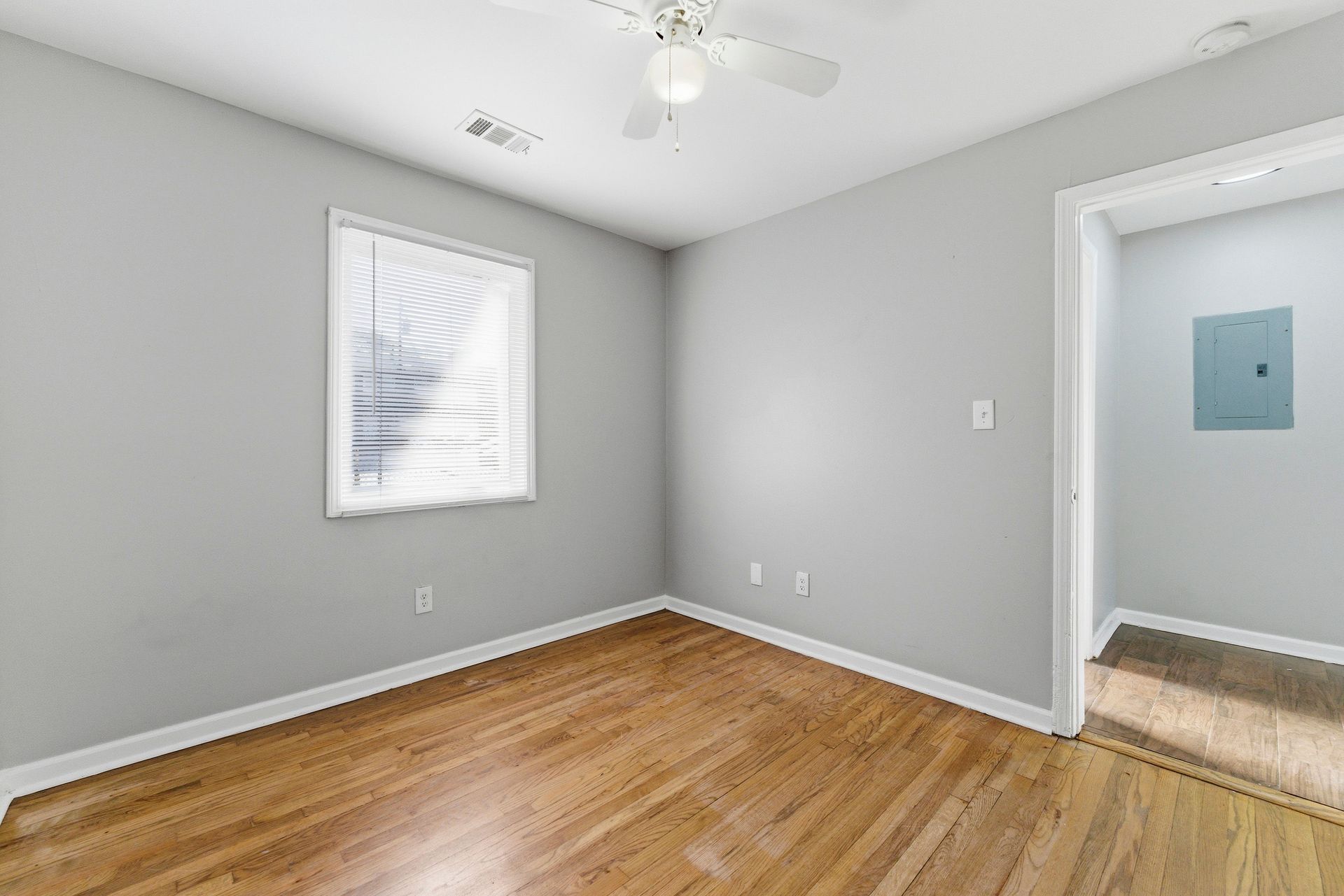 Empty gray bedroom with hardwood floors, a window, ceiling fan, and doorway to a carpeted hall