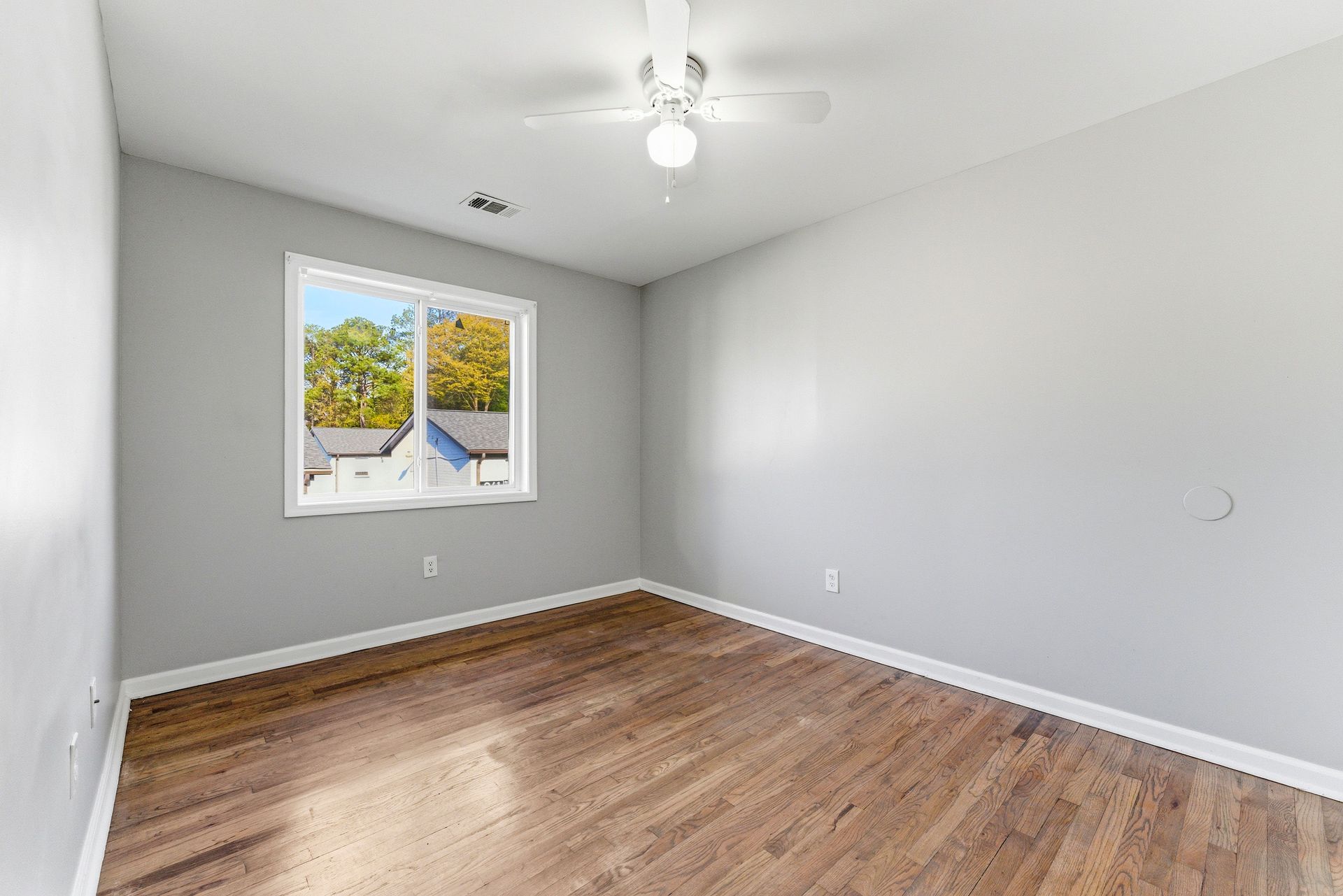 Empty gray bedroom with wood floors, a ceiling fan, and one window overlooking trees.