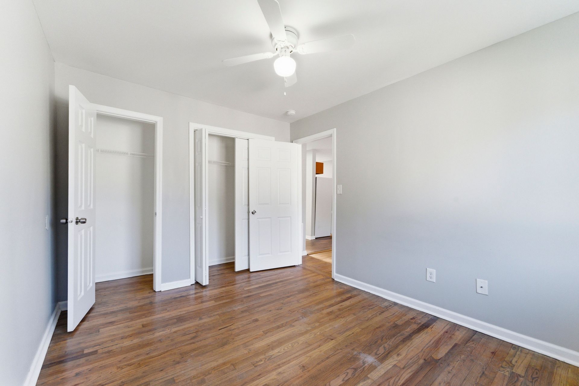 Empty white bedroom with hardwood floors, open closet doors, and a ceiling fan.