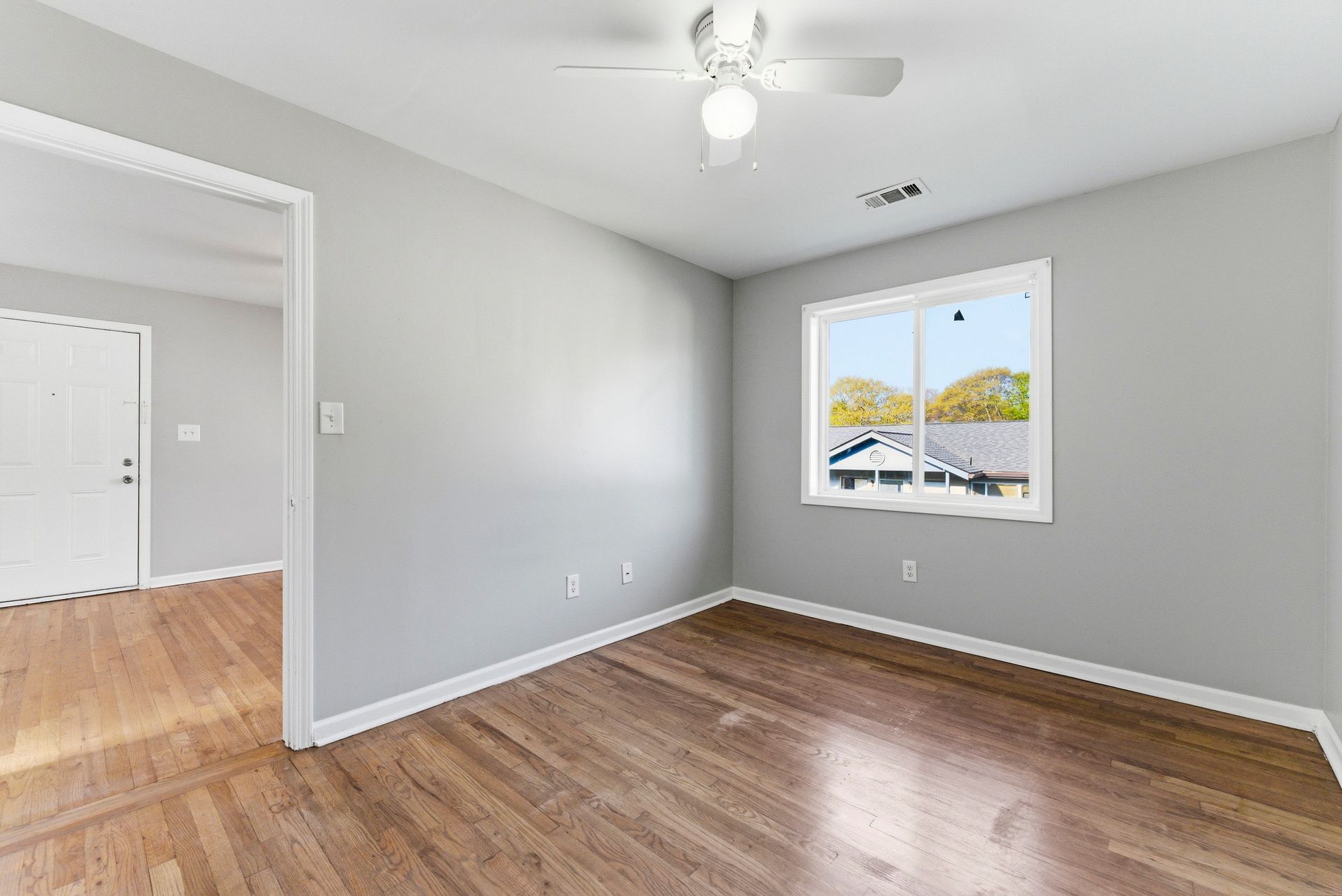 Empty gray bedroom with wood floors, ceiling fan, and a window overlooking trees