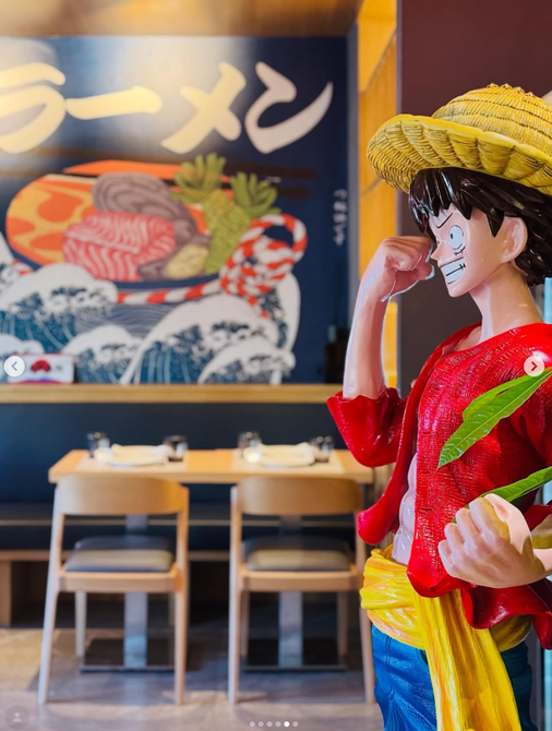 Figurina di un personaggio anime con camicia rossa e cappello di paglia all'interno di un ristorante di ramen con insegna giapponese.