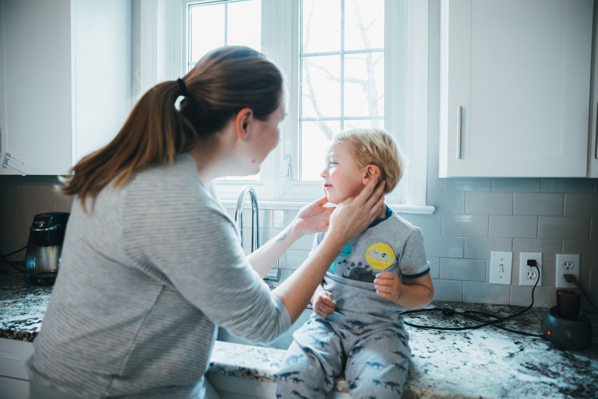 A woman is putting a pacifier in a baby's mouth while sitting on a kitchen counter.