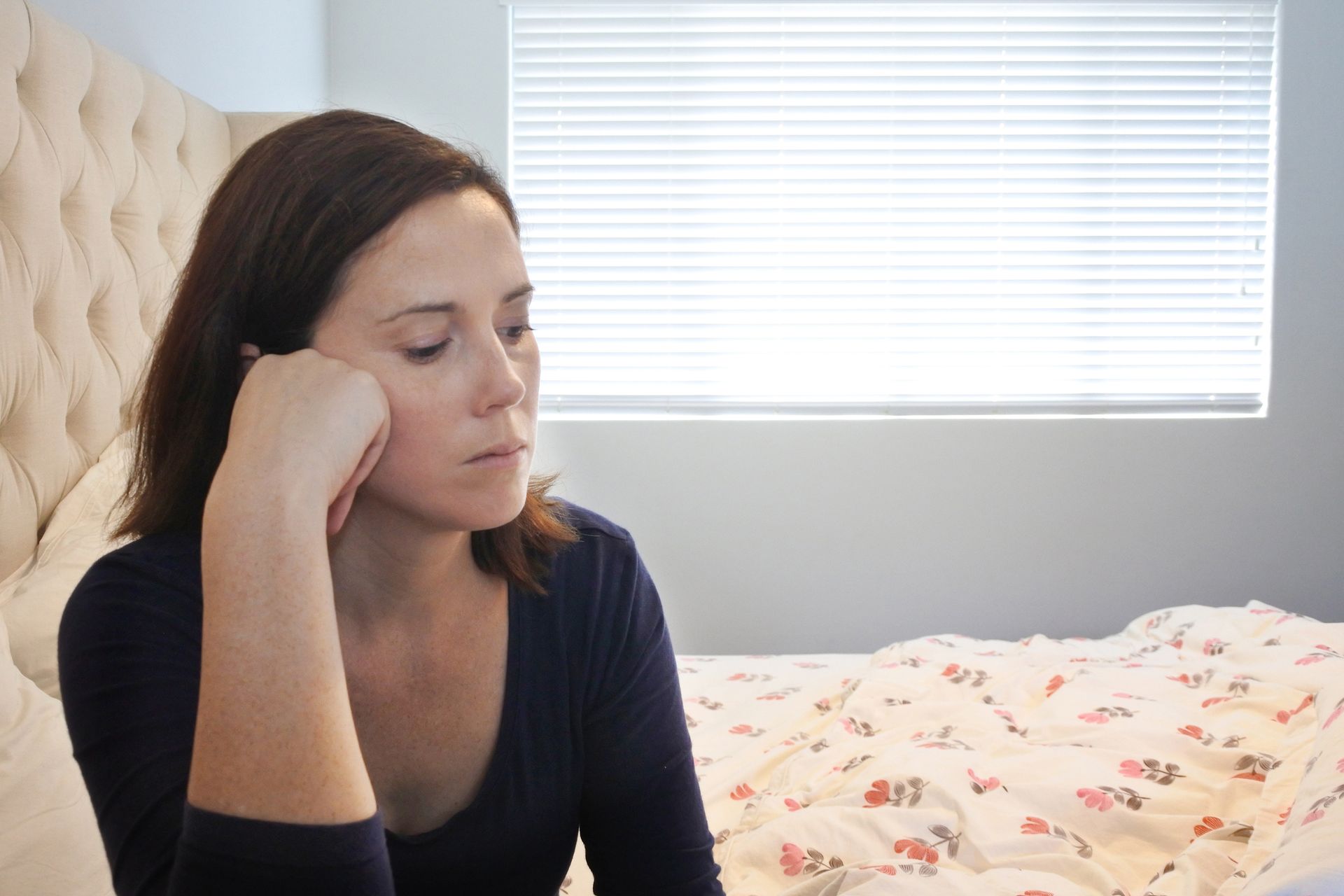 A woman is sitting on a bed with her head resting on her hand.