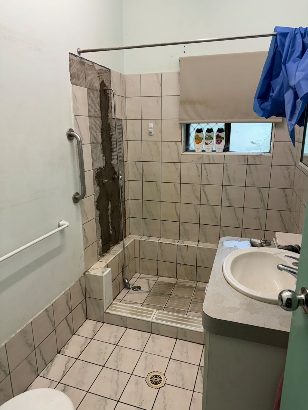 Small tiled bathroom shower with a damaged corner wall, sink, and blue towel hanging nearby — Nightcliff Plumbing & Gas In Nightcliff, NT