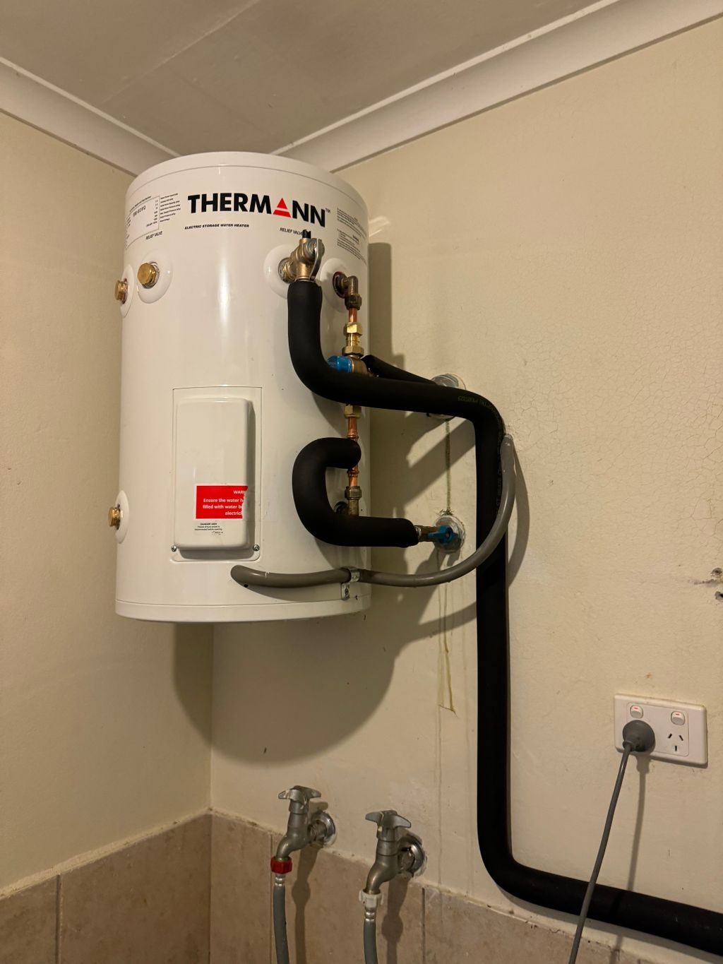 White wall-mounted water heater with black pipes in a brick-walled utility room — Nightcliff Plumbing & Gas In Coconut Grove, NT