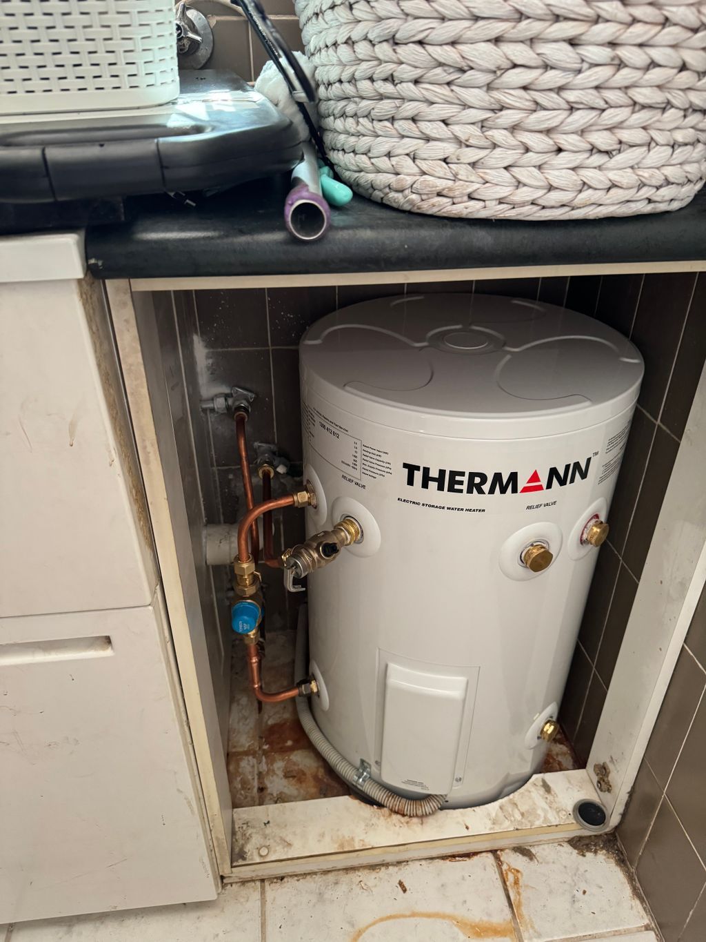 Thermann water heater in a small utility cabinet with pipes and a blue valve, partly blocked by stored items — Nightcliff Plumbing & Gas In Fannie Bay, NT