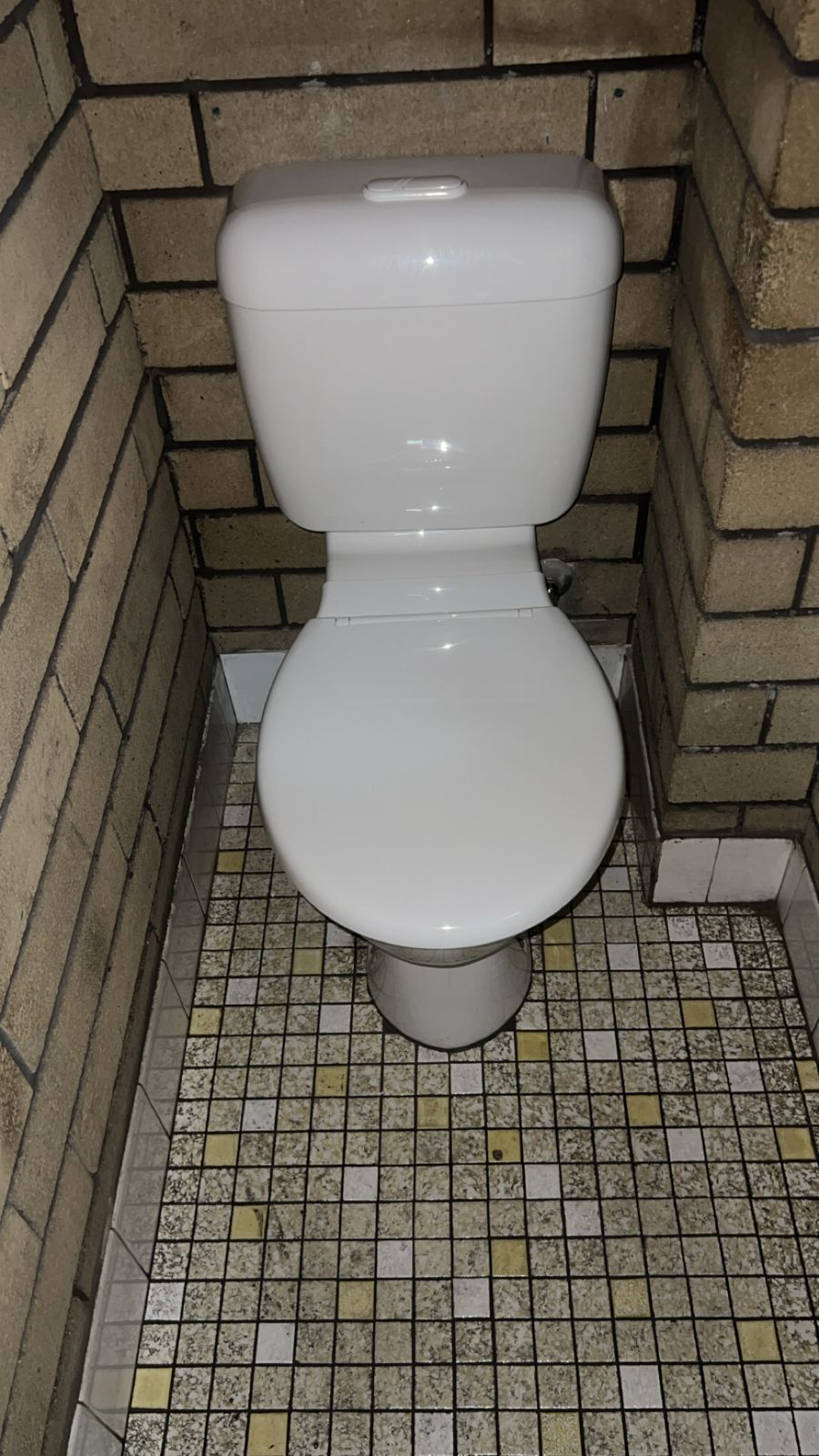 White toilet in a narrow tiled restroom stall with mosaic floor and brick-pattern walls — Nightcliff Plumbing & Gas In Stuart Park, NT