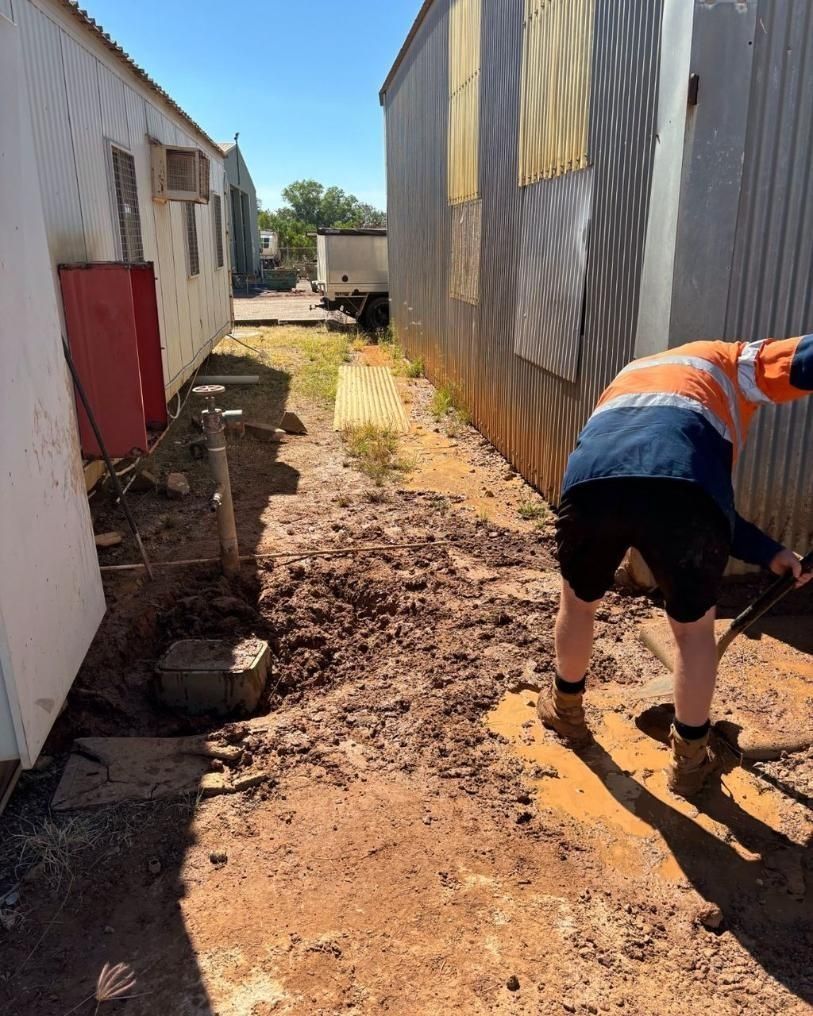 A Man is Digging in the Dirt Next to a Building With a Shovel — Nightcliff Plumbing & Gas In Humpty Doo, NT