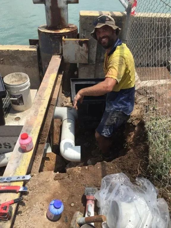 A Man is Kneeling in the Dirt Working on a Pipe — Nightcliff Plumbing & Gas In Coconut Grove, NT