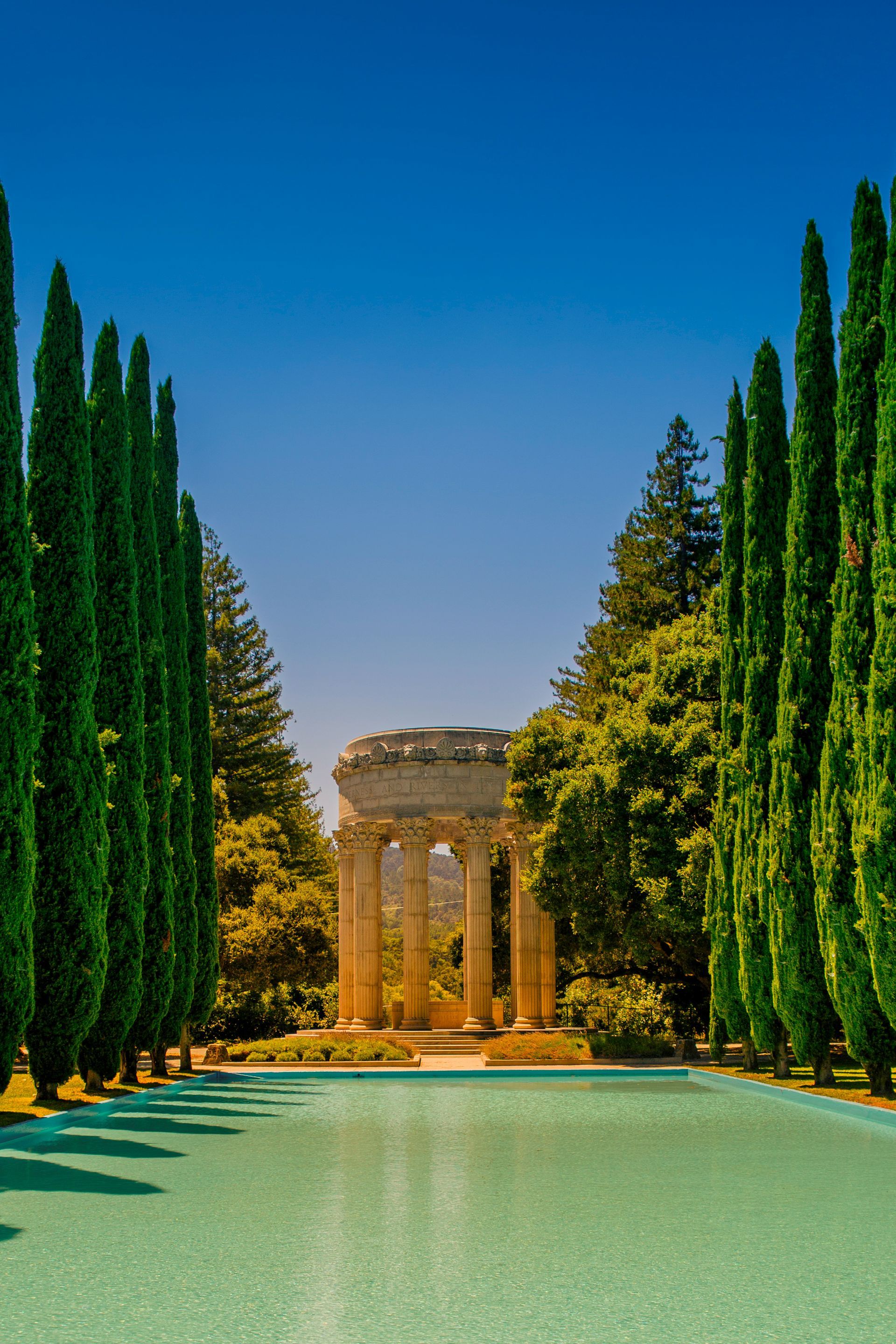 Tree-lined garden path with turquoise pool leading to a classical stone pavilion under a clear blue sky