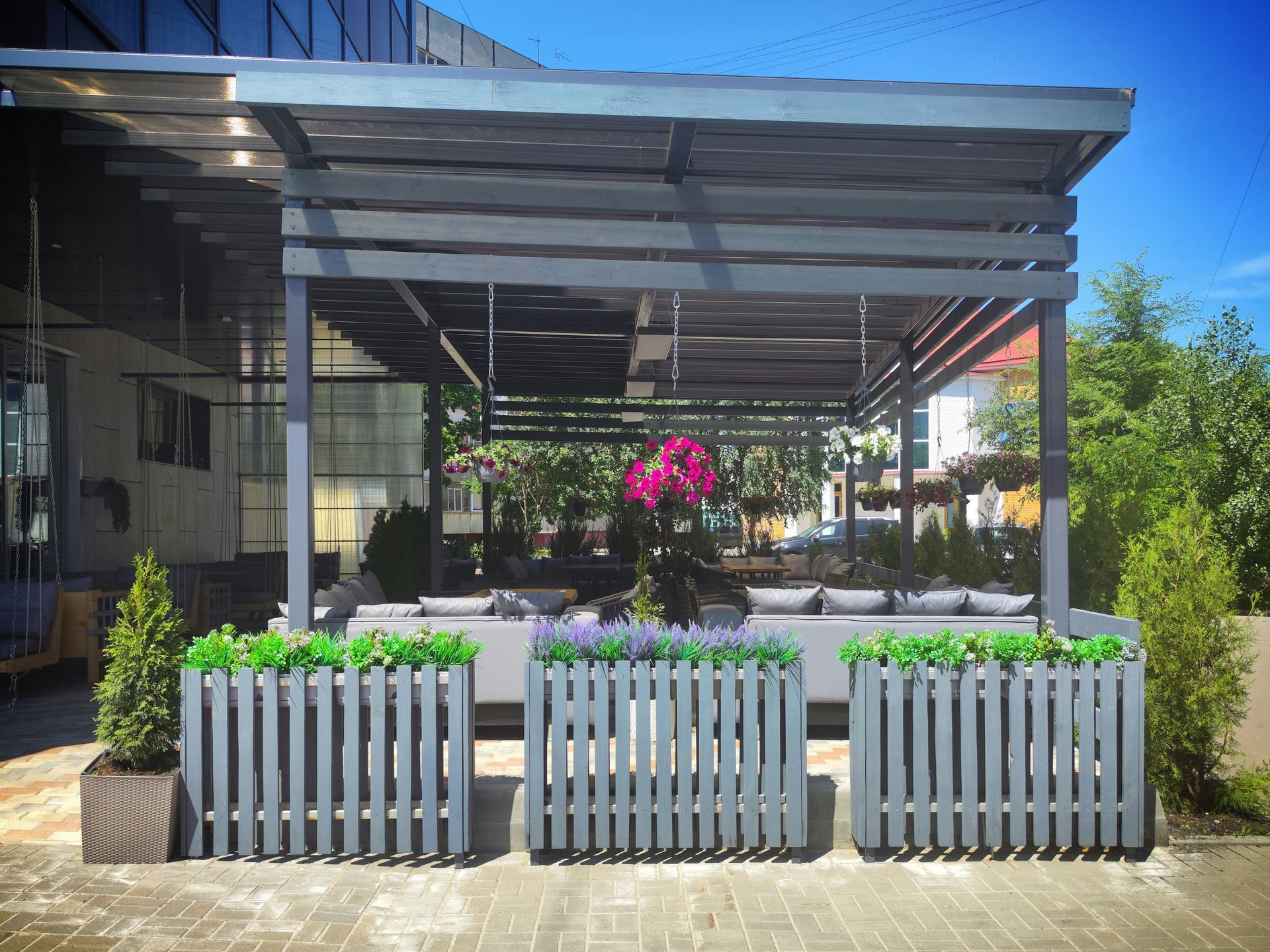 Outdoor patio with white picket planters, pink flowers, and a shaded metal pergola beside a building
