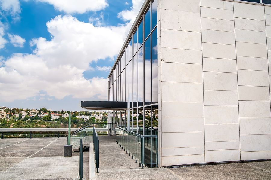 A modern building with a glass facade and stone wall, featuring an entrance awning and an adjacent paved parking area.