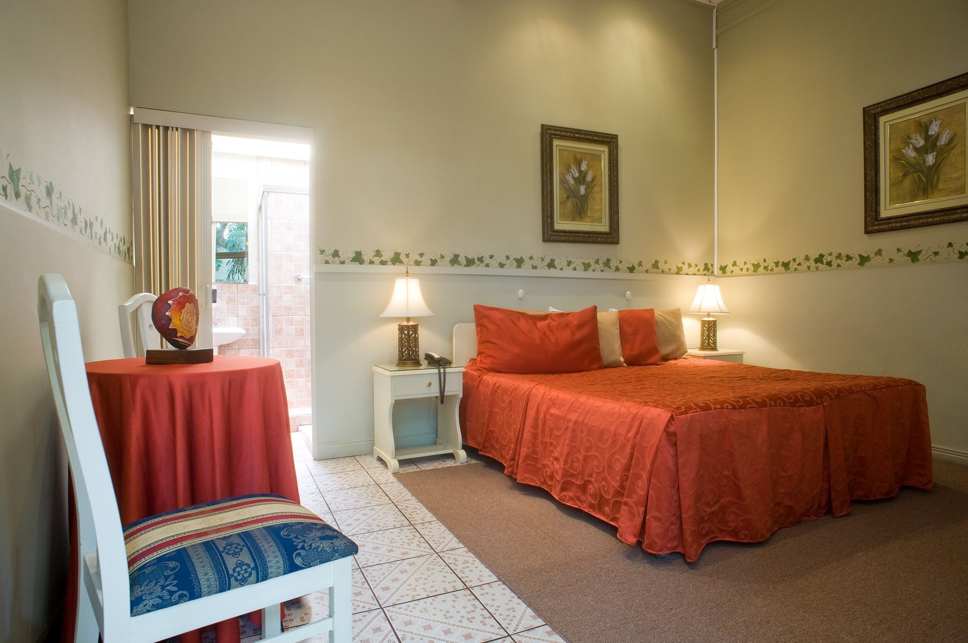 Cozy bedroom with red bedspread, chair, nightstand lamp, and sunlit window.