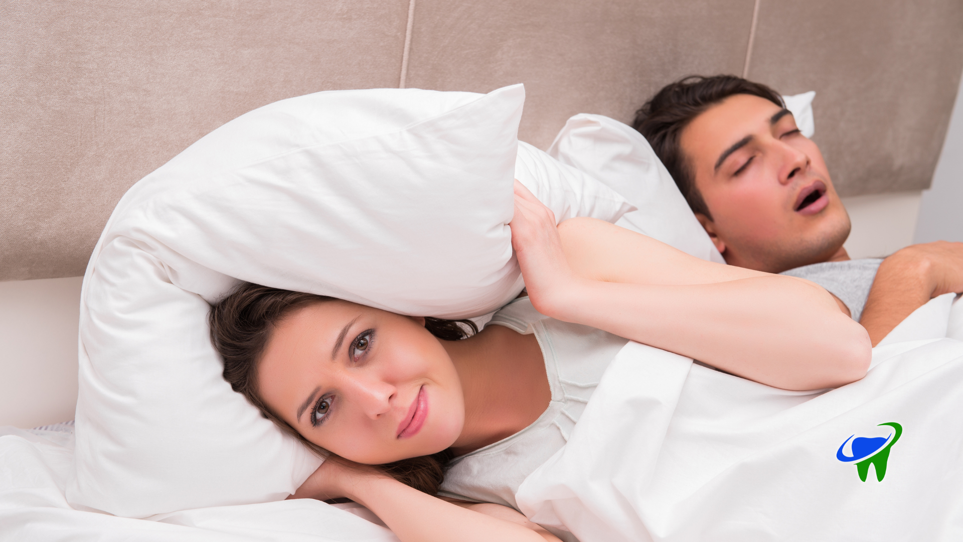 Woman lying in bed with a pillow over a sleeping man beside her, both under white bedding.