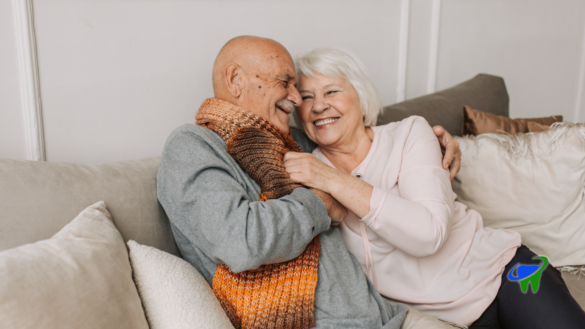 Two people smiling and hugging on a couch with white pillows, one holding an orange knit blanket