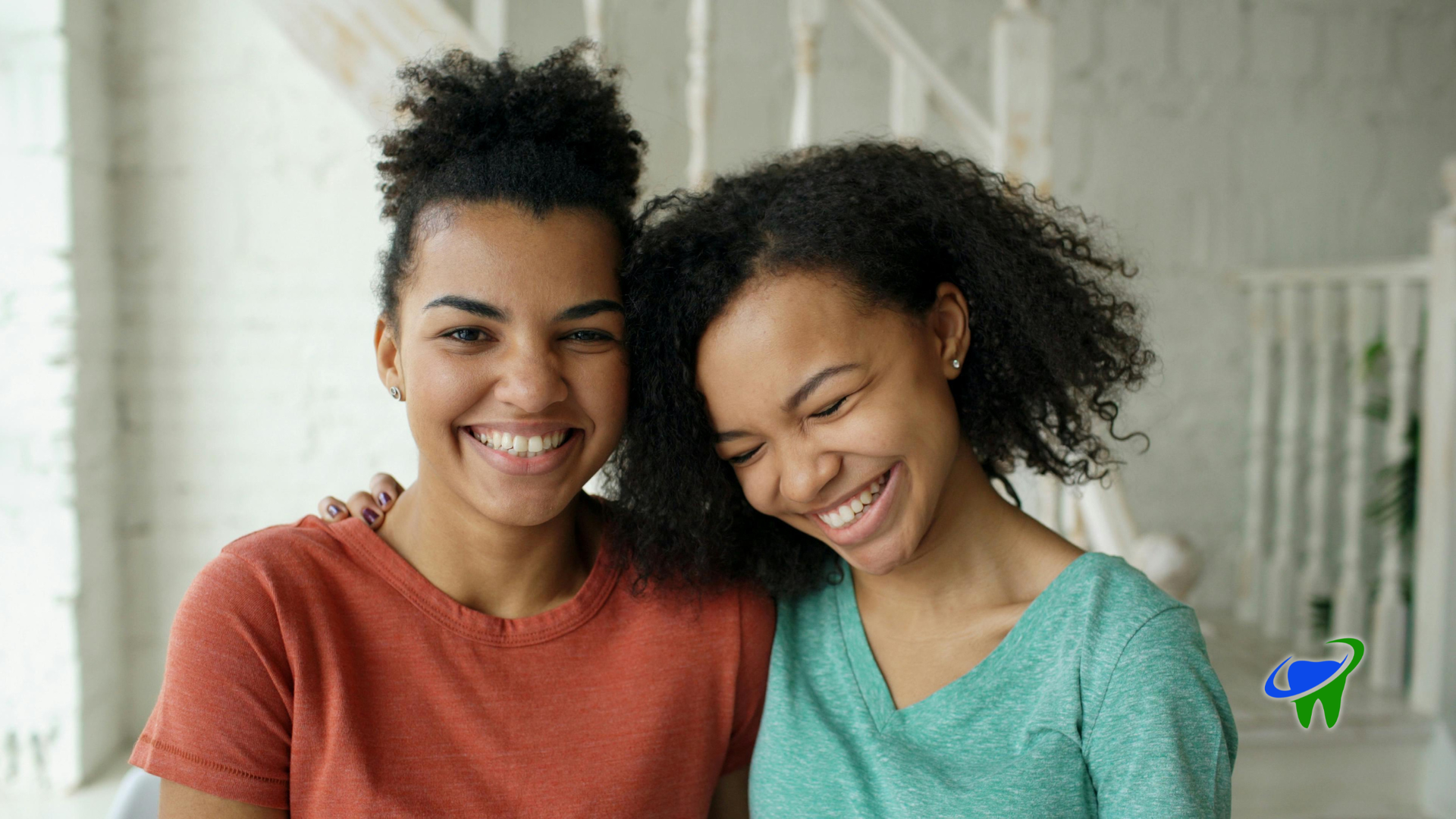 Two smiling people leaning together indoors, wearing orange and green tops.