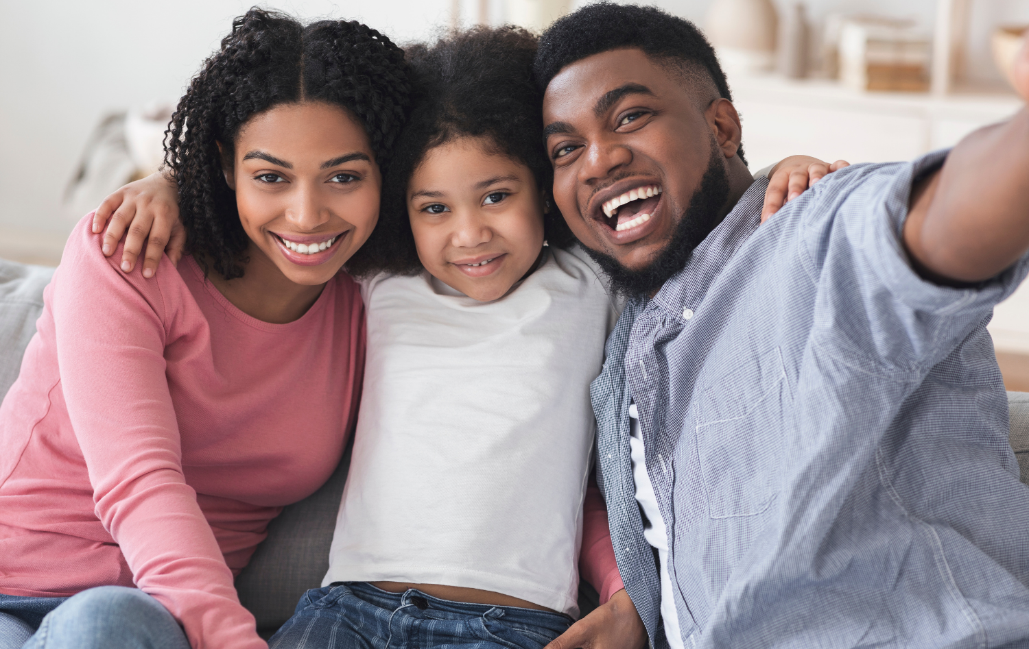 Three smiling people sitting close together on a couch, taking a selfie in a bright living room
