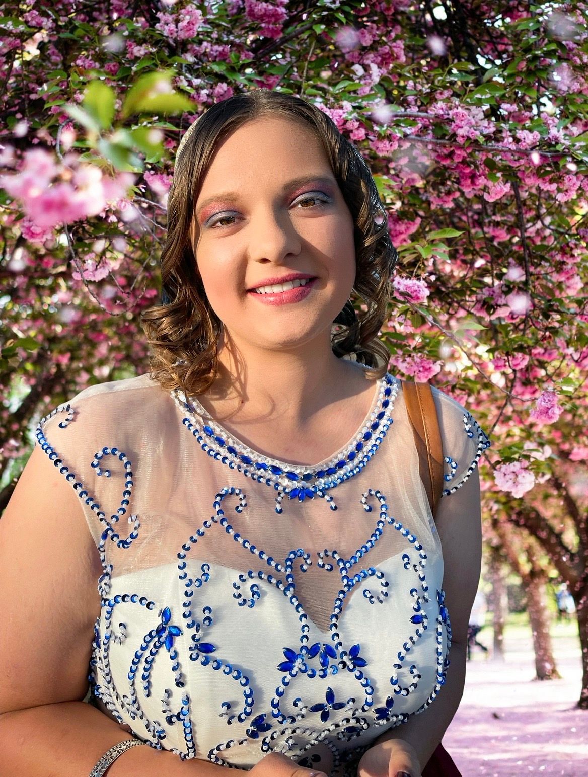 A person wearing a white dress with blue beading smiles while standing in front of blooming pink trees.