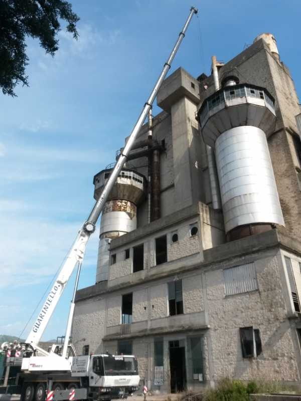 Una gru solleva un grande cilindro d'argento accanto a un edificio industriale in pietra, sotto un cielo azzurro.