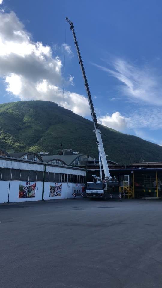 Un camion con gru solleva un lungo braccio accanto a un edificio, con montagne verdi e cielo azzurro sullo sfondo.