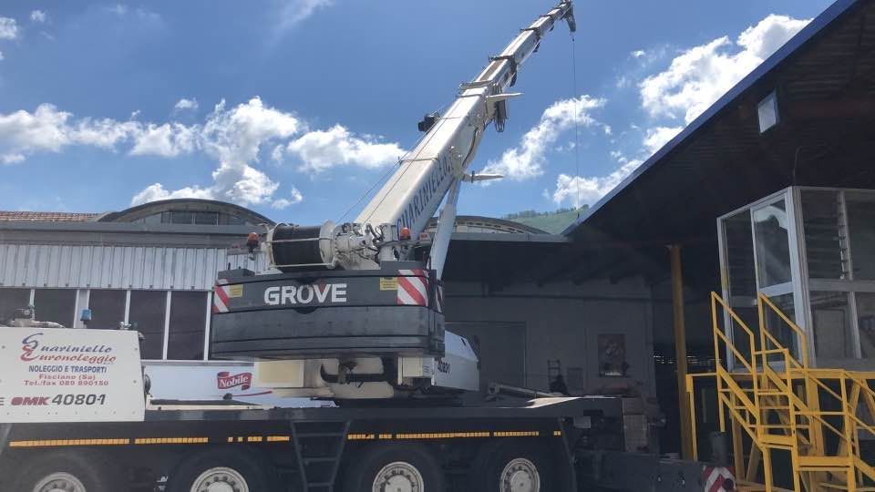 Una gru Grove su un camion accanto a un magazzino, con il braccio sollevato sotto un cielo azzurro.