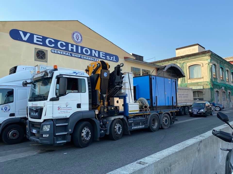 Carro attrezzi bianco che traina un rimorchio blu per attrezzature davanti a un edificio Vecchione su una strada