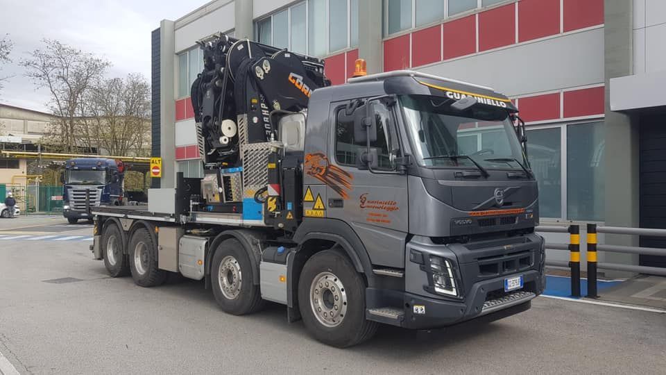 Un camion con gru grigio è parcheggiato davanti a un edificio, con una facciata rossa e bianca sullo sfondo.
