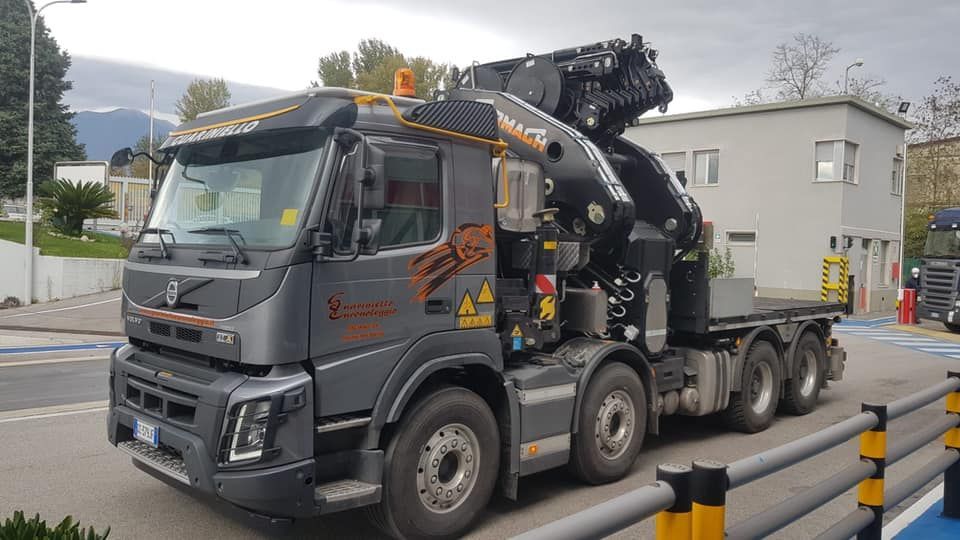 Camion grigio per carichi pesanti con gru parcheggiato su una strada vicino a un edificio e a delle barriere.