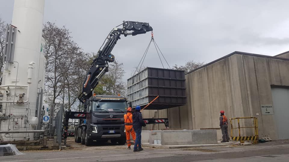 In un'area industriale, alcuni operai edili guidano una gru mentre solleva un grande container metallico.