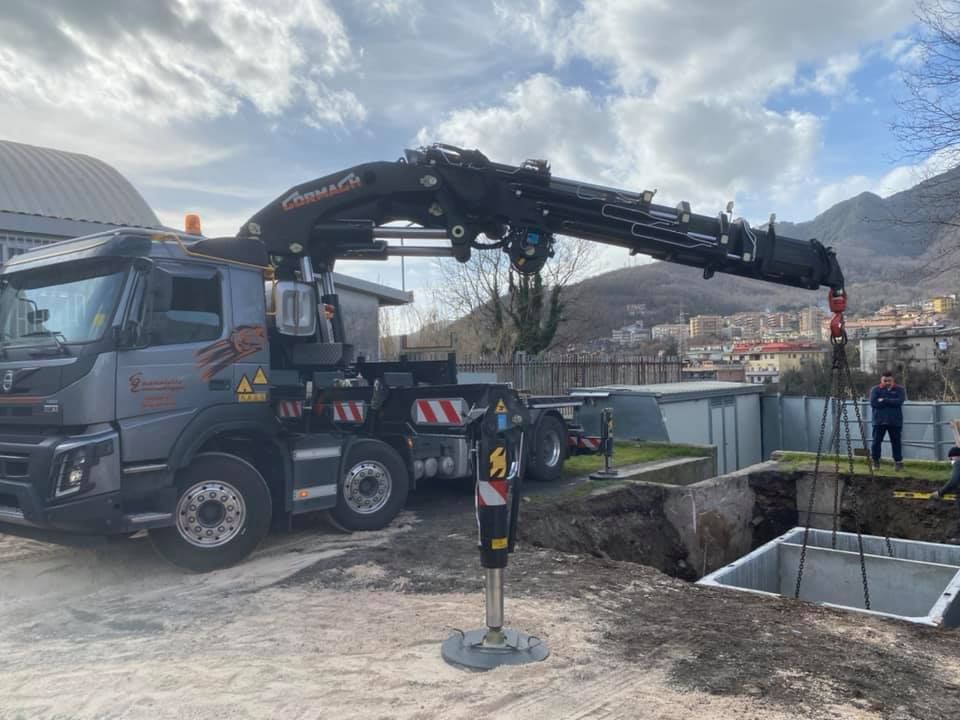Una gru montata su camion solleva un carico sopra uno scavo in un cantiere edile, con le montagne sullo sfondo.