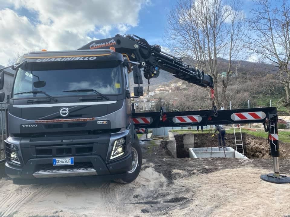 Carro attrezzi Volvo con pianale e braccio gru parcheggiato su una strada sterrata vicino a un ponte, con le montagne sullo sfondo.